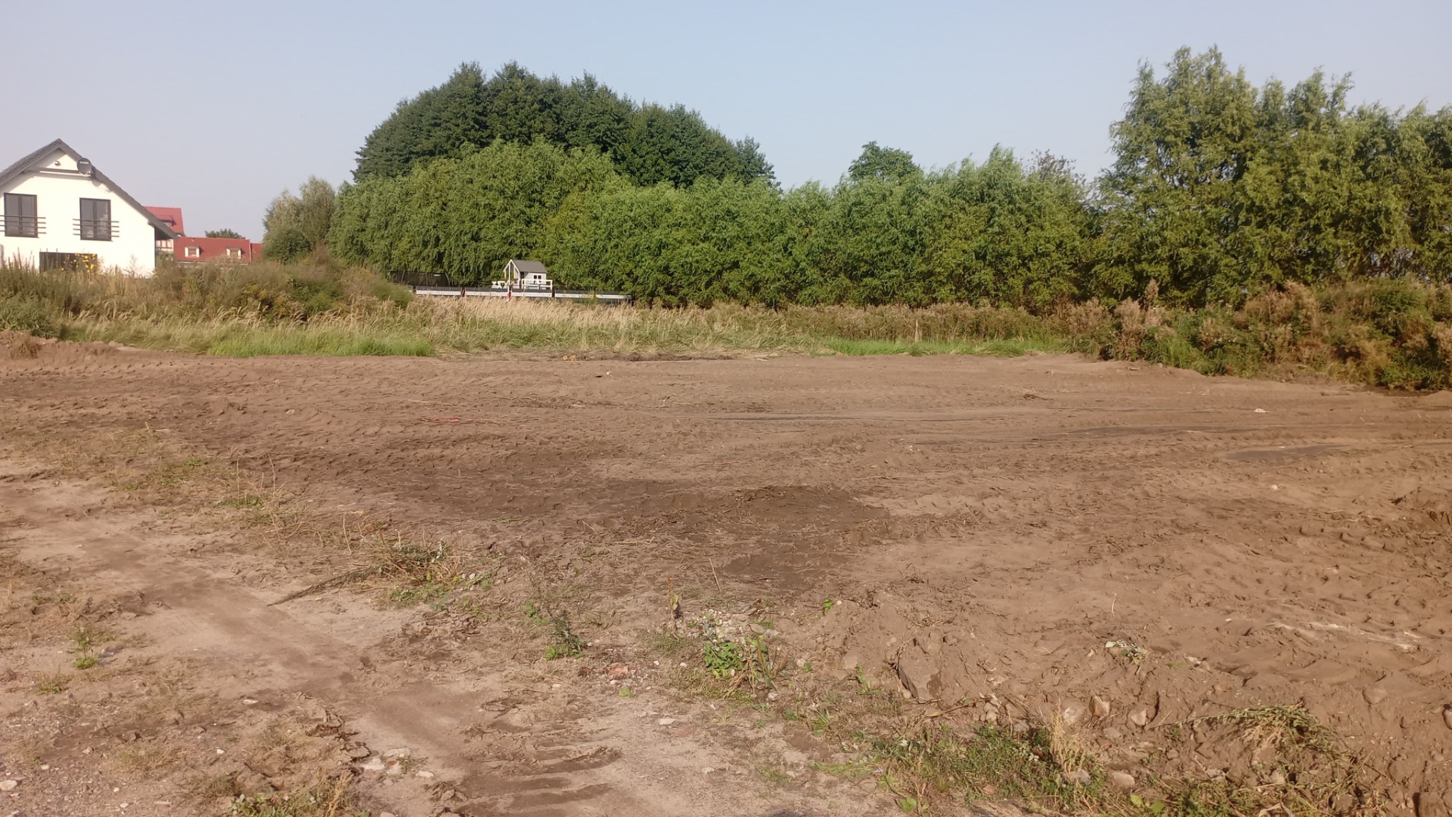 Niwelacja terenu pod budowę: wyrównana, brązowa ziemia na działce z widocznym domem i zielenią w tle, ślady opon maszyn budowlanych.