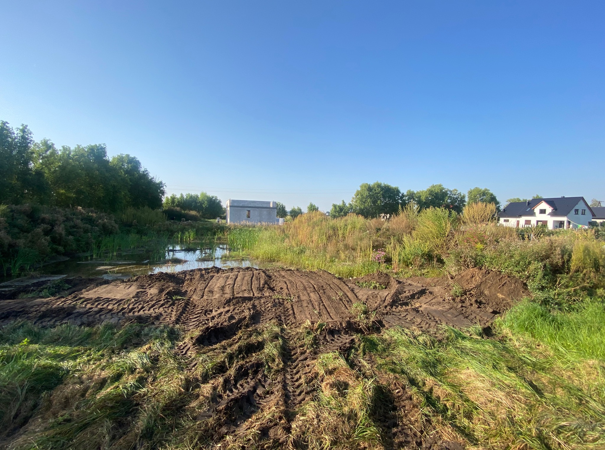 Krajobraz po niwelacji terenu z widocznymi śladami opon na ziemi, w tle zbiornik wodny, drzewa i budynki. Błękitne niebo nadaje świeżości.