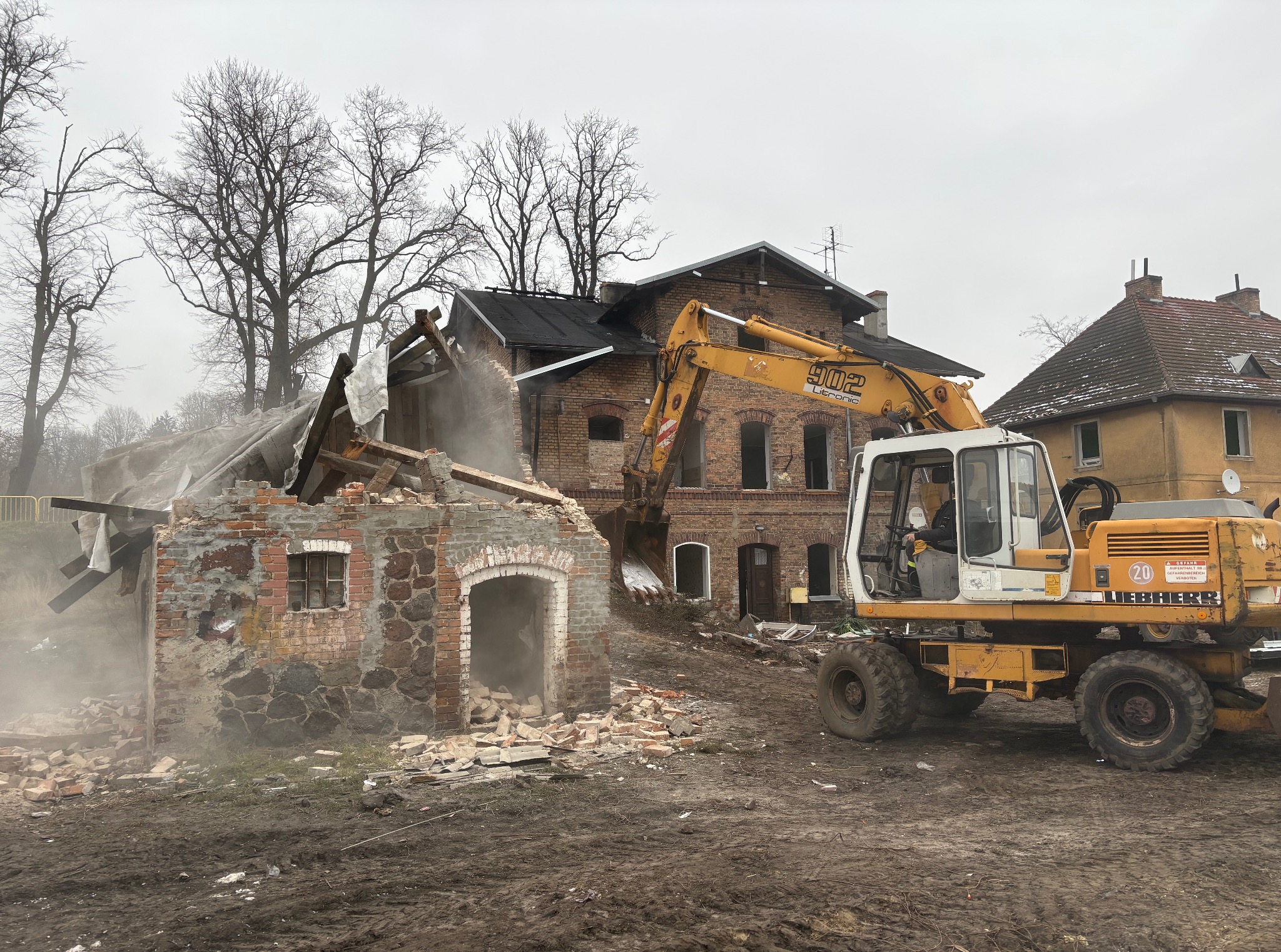 Rozbiórka ceglanego budynku w Drezdenko. Żółta koparka Liebherr w trakcie pracy, unoszący się pył i gruz. Częściowo zawalona konstrukcja.