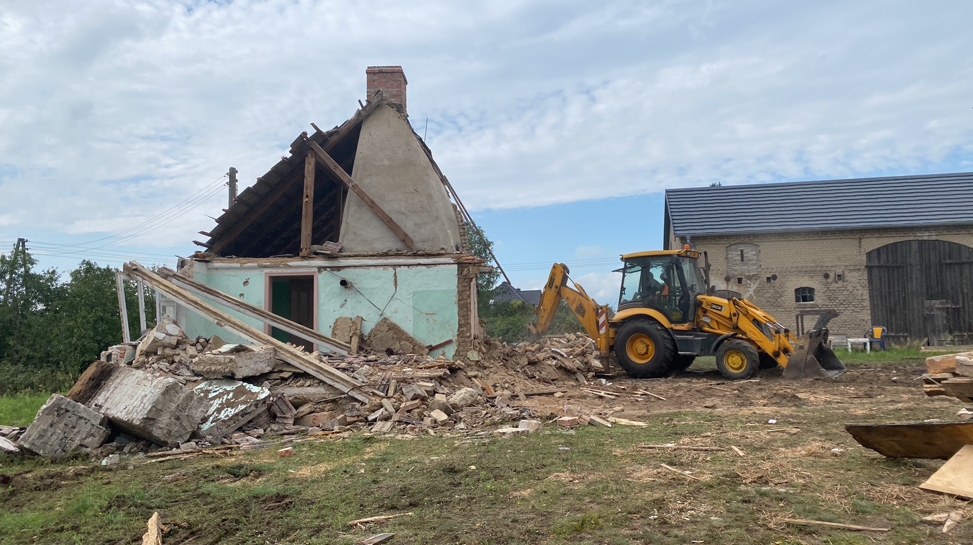 Żółta koparko-ładowarka JCB przy częściowo zburzonym, zielonym budynku z ceglanym kominem i stertą gruzu. W tle budynek gospodarczy z ciemnymi wrotami.