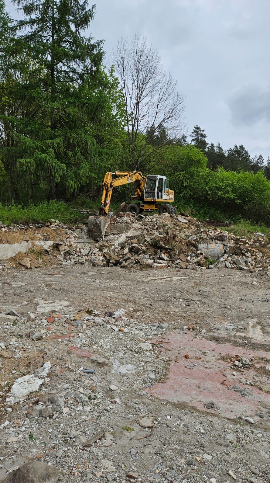 Żółta koparka na gruzowisku po rozbiórce budynku w otoczeniu drzew, pochmurny dzień. Widoczne fragmenty fundamentów i cegieł.