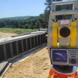 Żółto-szary teodolit na statywie mierzy betonową konstrukcję mostu na tle zieleni. Prace pomiarowe w terenie, budowa infrastruktury.