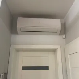 Biała klimatyzacja Electrolux zamontowana nad wejściem do pokoju, widoczny czujnik ruchu na ścianie. Minimalistyczny design wnętrza.