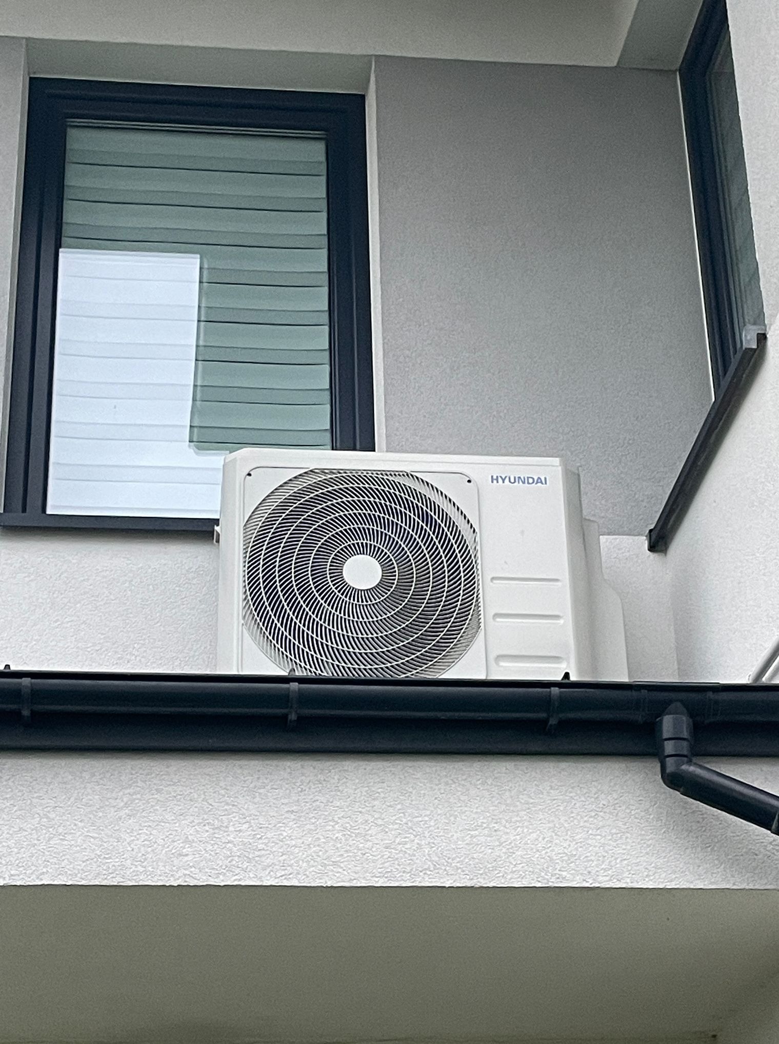 Biała jednostka zewnętrzna klimatyzacji Hyundai zamontowana na ścianie budynku, widoczna z boku, z czarną rynną i oknem z żaluzjami w tle.