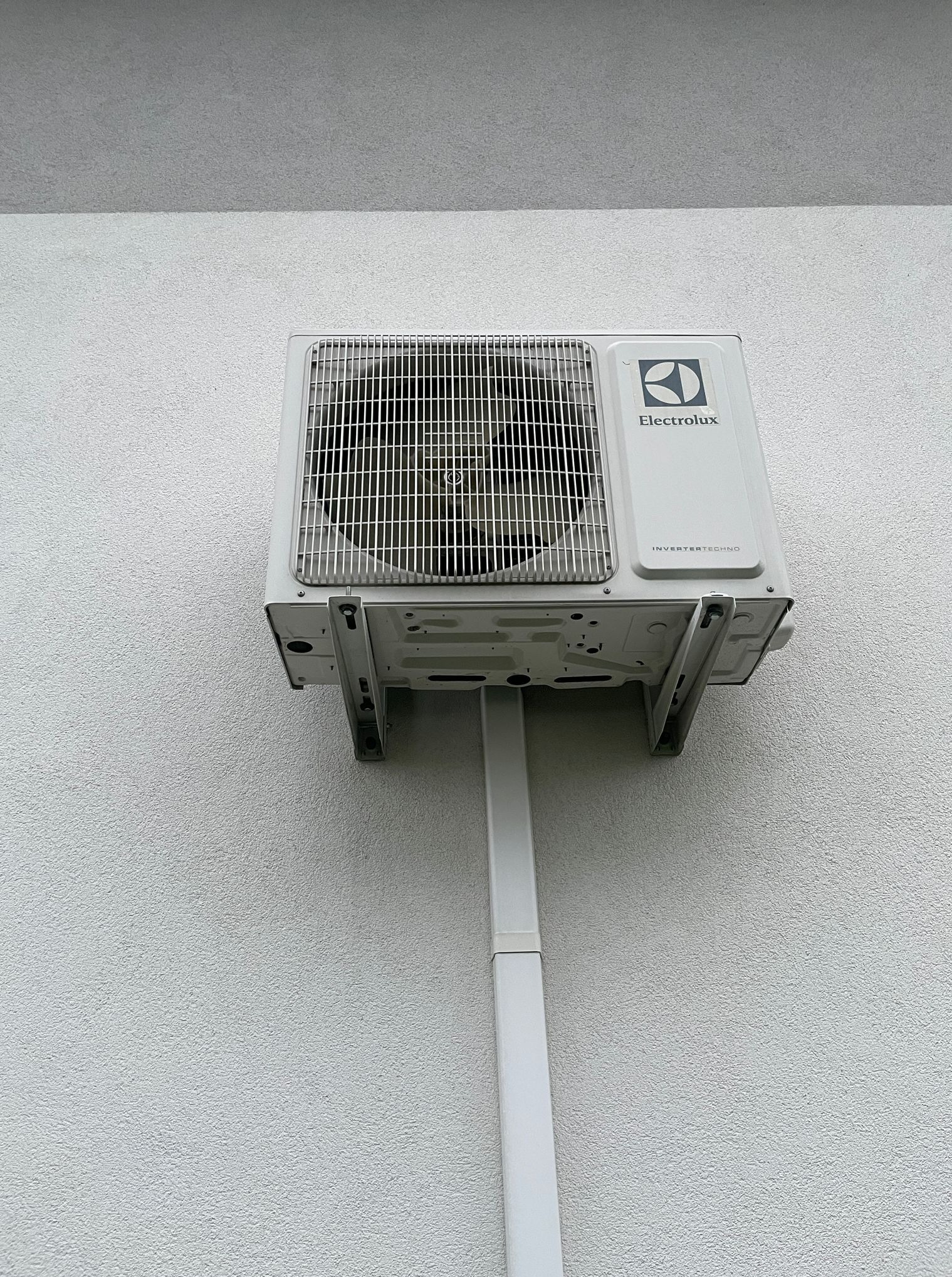 Biała jednostka zewnętrzna klimatyzacji Electrolux zamontowana na teksturowanej, jasnej ścianie budynku z widoczną rurą odprowadzającą skropliny.