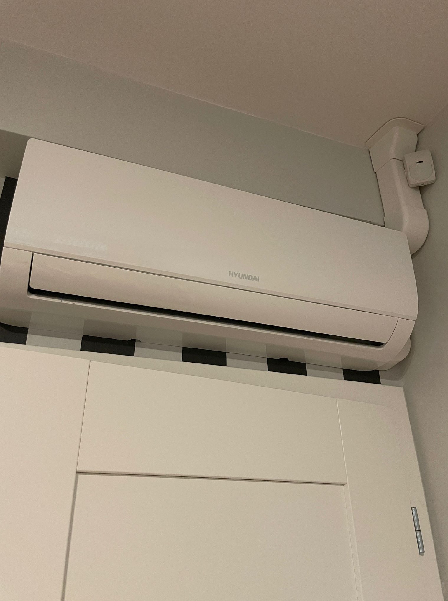 Biała klimatyzacja Hyundai na ścianie nad szafką, z widoczną instalacją odprowadzającą. Ujęcie z dołu, widoczne elementy wnętrza.