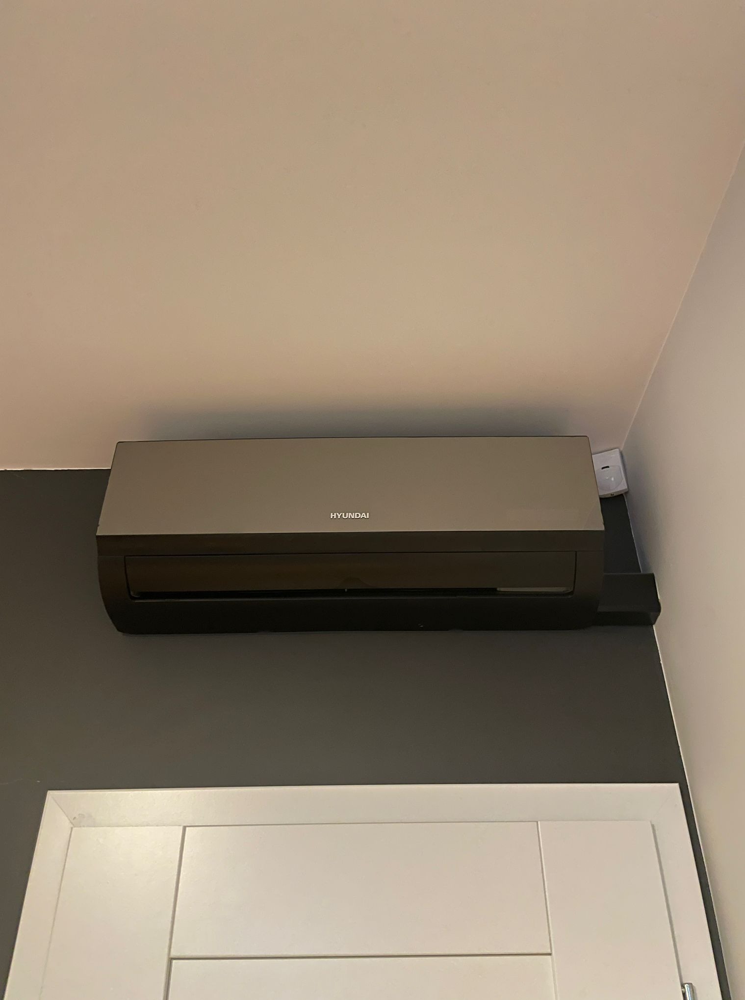 Szary klimatyzator Hyundai, zamontowany na ciemnej ścianie nad białą futryną drzwiową. Widoczne gniazdko elektryczne obok urządzenia.