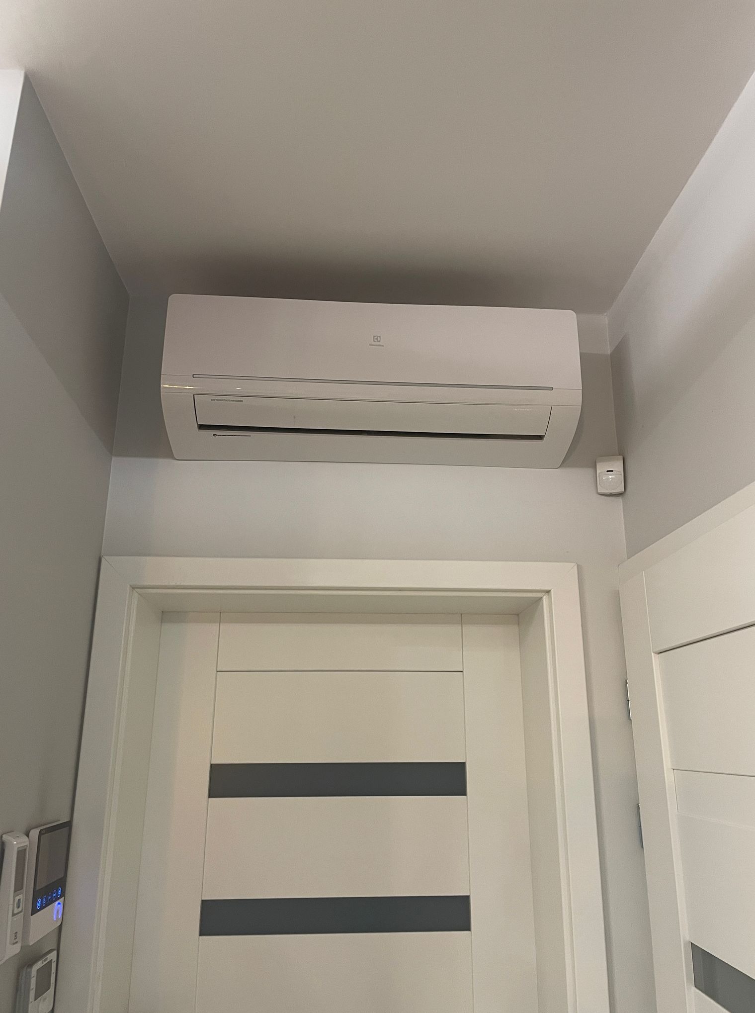 Biała klimatyzacja Electrolux zamontowana nad wejściem do pokoju, widoczny czujnik ruchu na ścianie. Minimalistyczny design wnętrza.