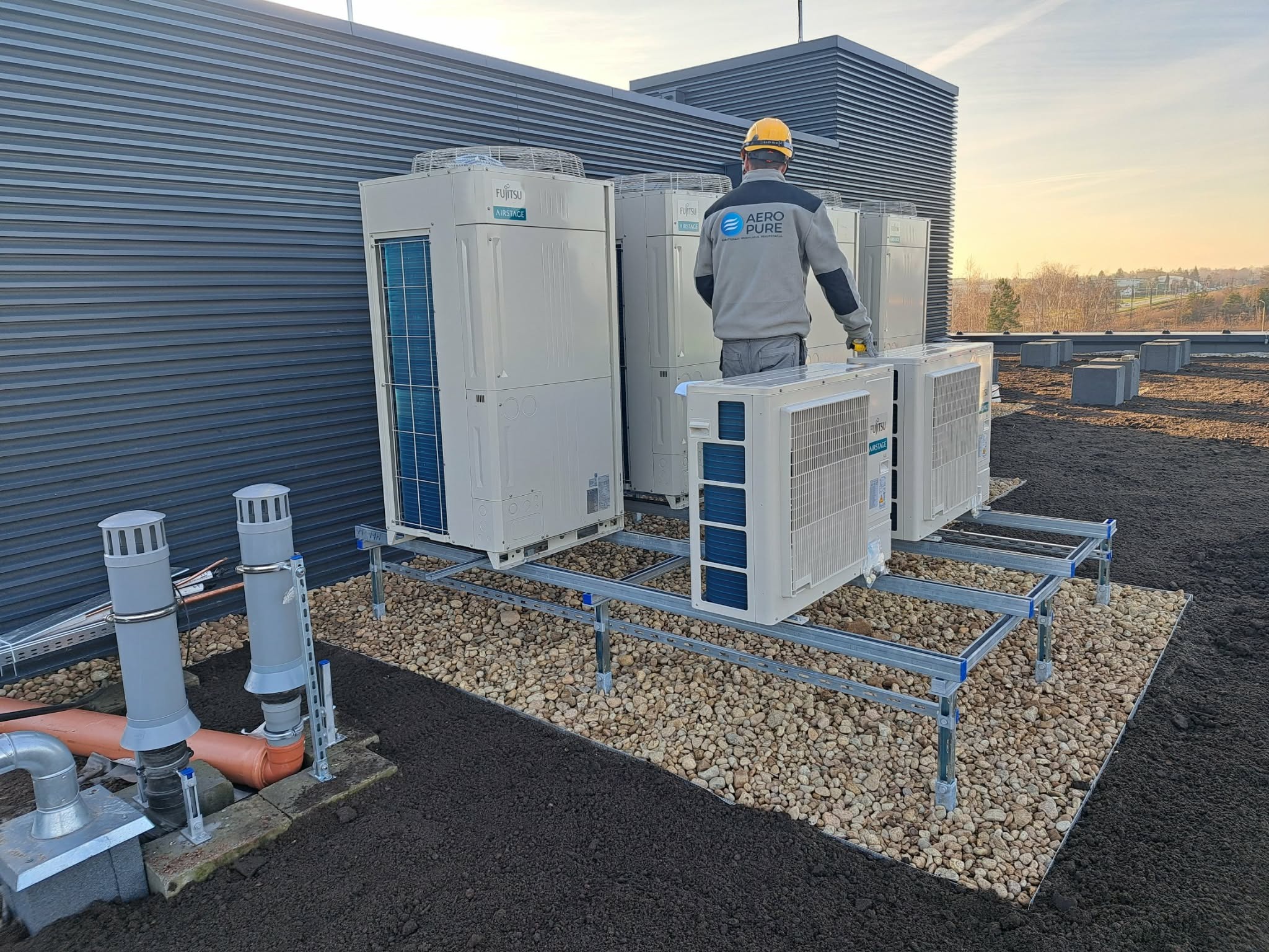 Instalacja klimatyzacji Fujitsu Airstage na dachu budynku komercyjnego. Widoczny monter w kasku i firmowej bluzie Aero Pure. Tło stanowi panorama miasta.