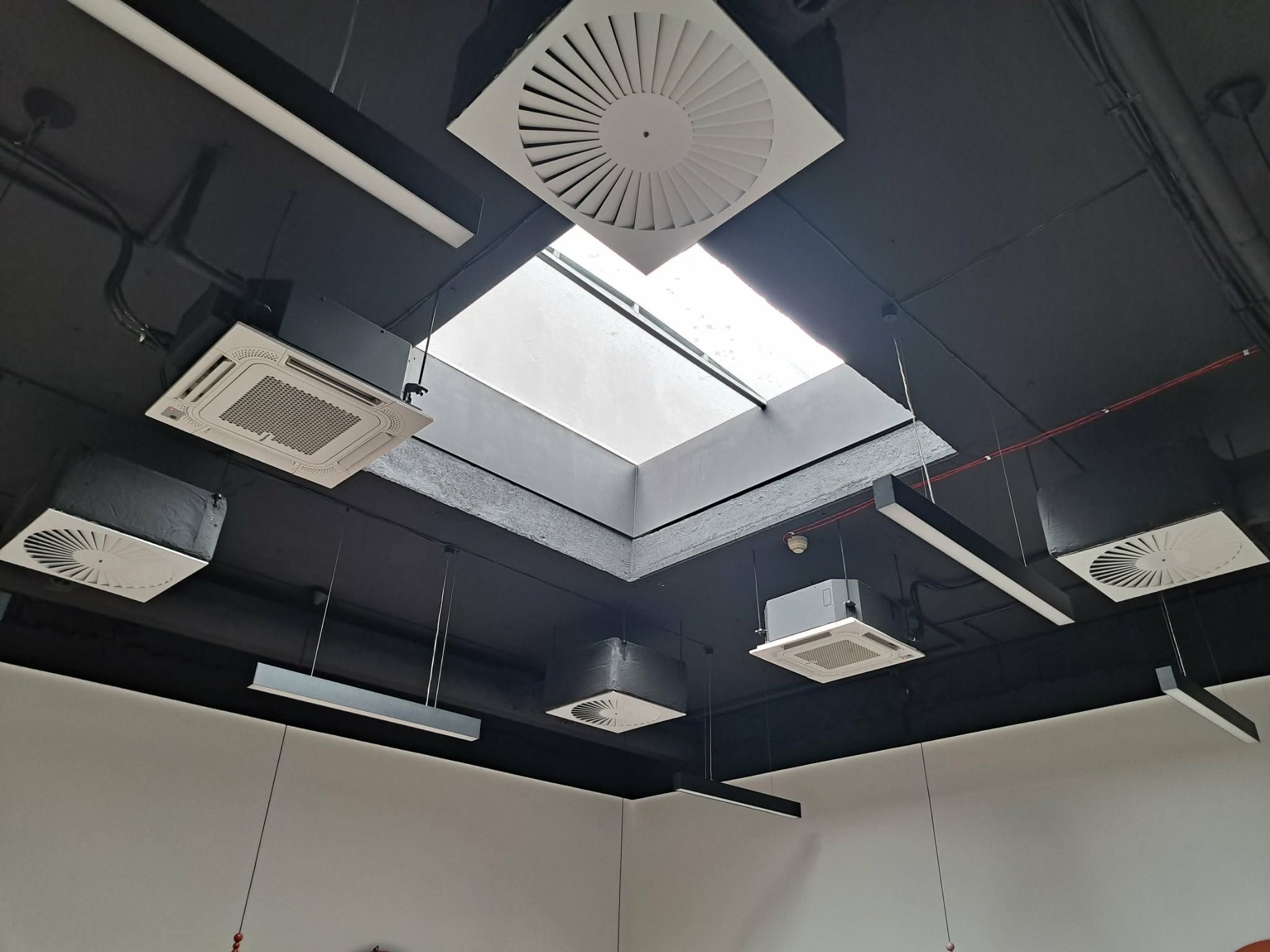 Czarny sufit z zamontowanymi białymi klimatyzatorami kasetonowymi i okrągłymi anemostatami, oświetlony przez podłużne lampy i naturalne światło ze świetlika.