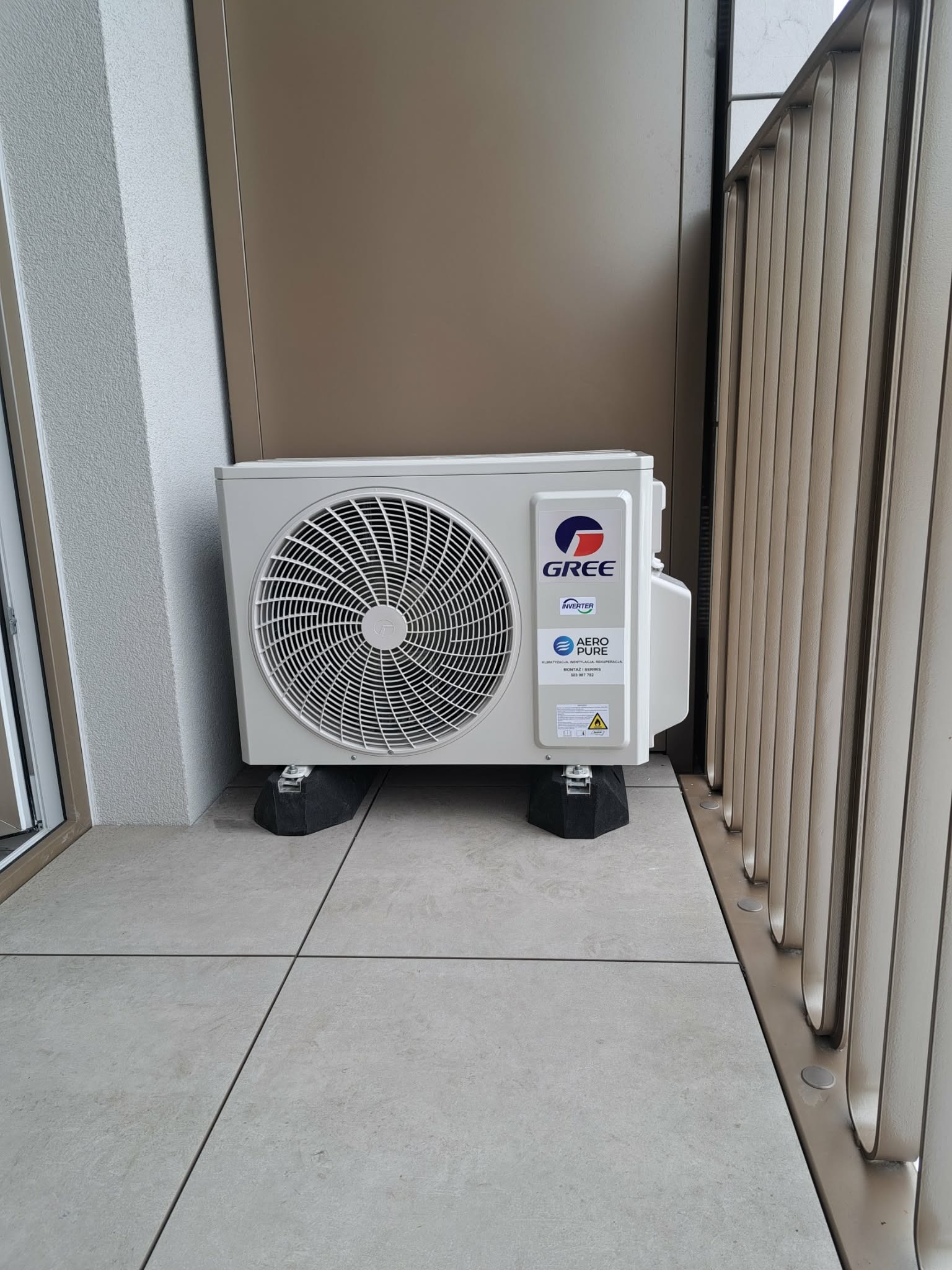 Biała jednostka zewnętrzna klimatyzacji marki Gree, umieszczona na czarnych podkładkach antywibracyjnych na balkonie z szarą podłogą i metalową balustradą.