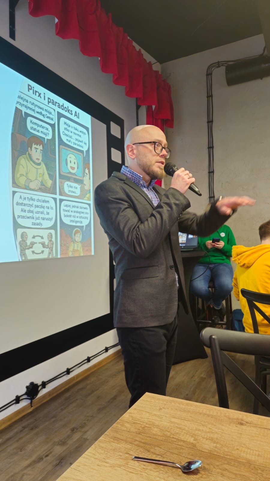 Prelegent w szarej marynarce prezentuje slajd o 'Pirxie i paradoksach AI', z komiksowymi kadrami. W tle czerwone zasłony i fragment sali konferencyjnej.