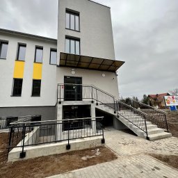 DOLIŃSKI BUDOWNICTWO DAWID DOLIŃSKI - Nowoczesny budynek z rampą i schodami wejściowymi, szara elewacja z żółtymi akcentami, czarne balustrady, kostka brukowa na podejściu, pochmurne niebo w tle.