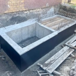 DOLIŃSKI BUDOWNICTWO DAWID DOLIŃSKI - Zabezpieczony, betonowy fundament z wystającymi prętami zbrojeniowymi, częściowo zakryty deskami. Widoczna izolacja przeciwwilgociowa. Prace ziemne w toku.