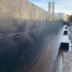 DOLIŃSKI BUDOWNICTWO DAWID DOLIŃSKI - Zabezpieczona, czarną izolacją pionowa ściana fundamentowa z betonu, z widocznymi prętami zbrojeniowymi i wykopem. Świeżo wykonana hydroizolacja.