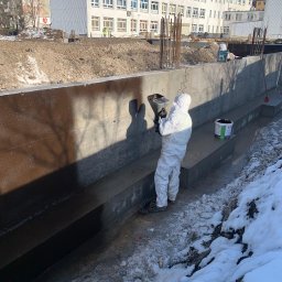 DOLIŃSKI BUDOWNICTWO DAWID DOLIŃSKI - Pracownik w kombinezonie ochronnym aplikuje hydroizolację na betonową ścianę fundamentową. Widoczny sprzęt do natrysku i wiadro z preparatem. W tle wykop i budynek.