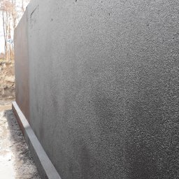 DOLIŃSKI BUDOWNICTWO DAWID DOLIŃSKI - Szara, teksturowana ściana fundamentowa z widoczną izolacją przeciwwodną i obrzeżem, na tle ziemi i drzew bez liści.