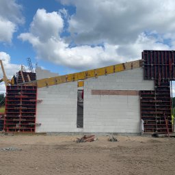 DOLIŃSKI BUDOWNICTWO DAWID DOLIŃSKI - Wznoszenie ścian domu z białych bloczków, z użyciem szalunków systemowych i dźwigu teleskopowego JCB na tle błękitnego nieba z chmurami.