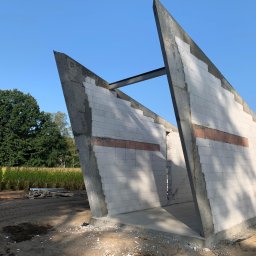 DOLIŃSKI BUDOWNICTWO DAWID DOLIŃSKI - Surowe ściany z betonu komórkowego i żelbetu w nietypowym kształcie, z widoczną stalową belką, na tle błękitnego nieba i zieleni. Budowa w toku, widoczne materiały budowlane.