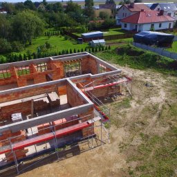DOLIŃSKI BUDOWNICTWO DAWID DOLIŃSKI - Wysoka perspektywa na budowę domu z czerwonej cegły, z widocznymi rusztowaniami i betonowymi nadprożami, na tle zielonego krajobrazu z domami.