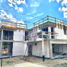 DOLIŃSKI BUDOWNICTWO DAWID DOLIŃSKI - Wznoszenie dwukondygnacyjnego domu z białych bloczków, z widocznym zbrojeniem i drewnianymi barierkami. Na górze baner reklamowy firmy budowlanej. Błękitne niebo w tle.