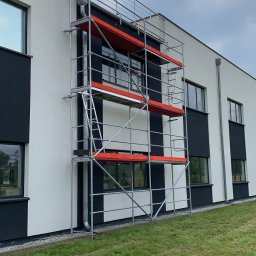 DOLIŃSKI BUDOWNICTWO DAWID DOLIŃSKI - Elewacja budynku z rusztowaniem na tle zielonego trawnika i pochmurnego nieba. Widoczne okna i elementy wykończeniowe w kolorze czarnym.