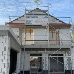 DOLIŃSKI BUDOWNICTWO DAWID DOLIŃSKI - Budowa domu z białych bloczków, widoczne rusztowania i baner firmy DD Budownictwo. Częściowo otynkowane ściany, okna bez szyb. W tle błękitne niebo.