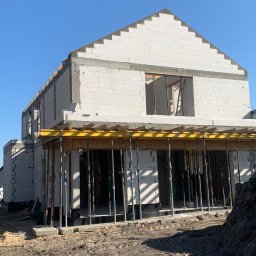 DOLIŃSKI BUDOWNICTWO DAWID DOLIŃSKI - Wznoszenie ścian z białych bloczków na budowie domu jednorodzinnego. Widoczne podpory stropu i ślady opon na ziemi. Słoneczny dzień, błękitne niebo.