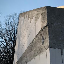 DOLIŃSKI BUDOWNICTWO DAWID DOLIŃSKI - Surowa ściana z betonu komórkowego w trakcie budowy, widoczne elementy konstrukcyjne i fragmenty drzew na tle jasnego nieba. Detal budowlany w zbliżeniu.