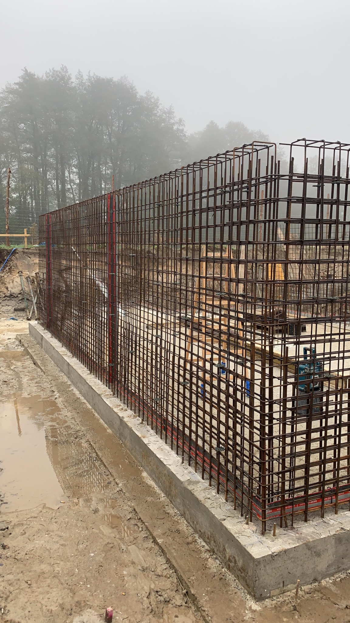 Zbrojenie fundamentu z betonu w szary dzień. Widoczne stalowe pręty tworzące siatkę, betonowa podstawa i błotniste podłoże. W tle zalesiony teren spowity mgłą.