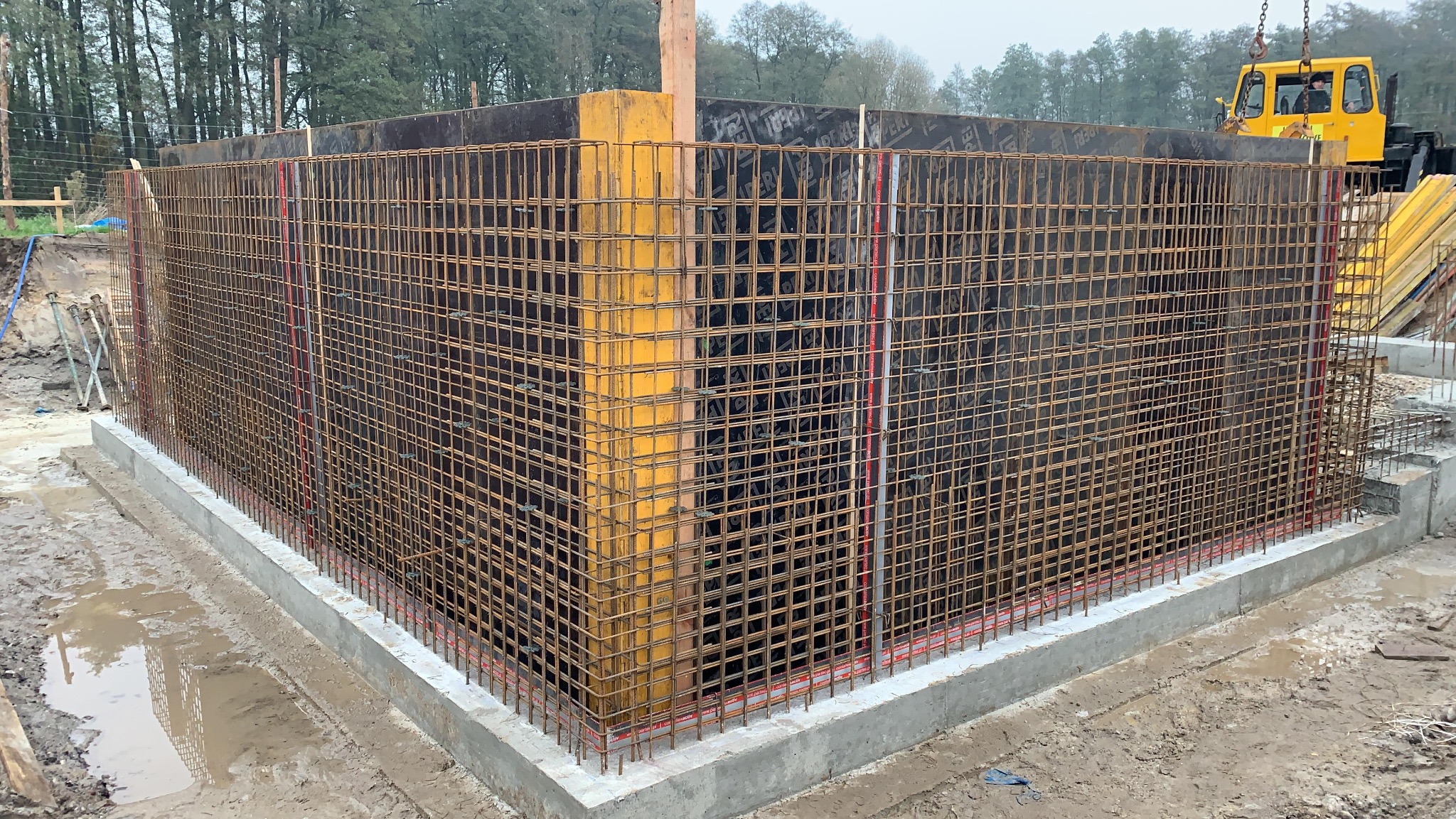 Zbrojenie fundamentu z szalunkiem i widocznym żółtym dźwigiem w tle. Siatka z prętów stalowych tworzy konstrukcję pod wylewkę betonową.