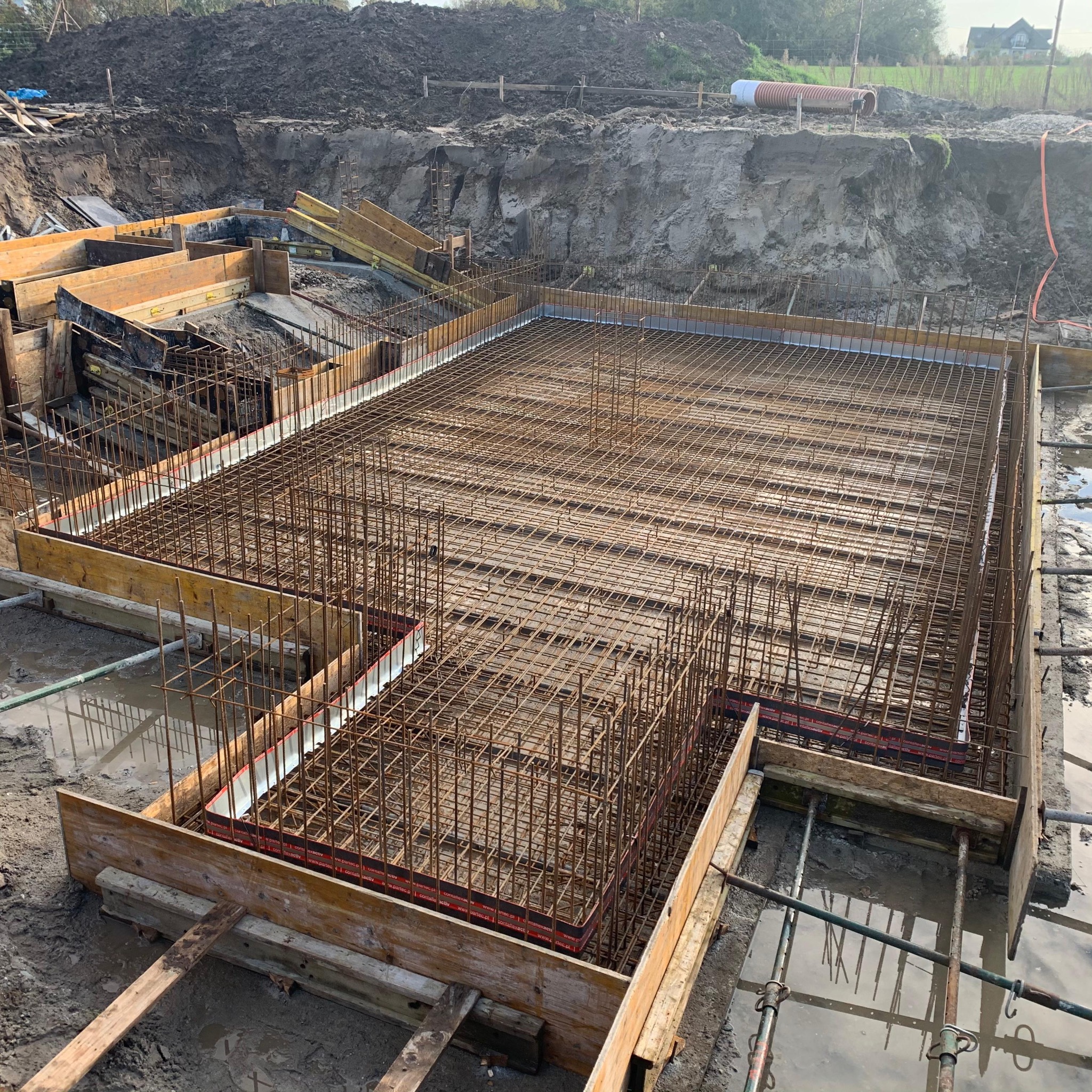 Zbrojenie fundamentu w drewnianym szalunku, widok z góry. Stalowe pręty tworzą siatkę, przygotowane do zalania betonem. W tle wykop i hałda ziemi.