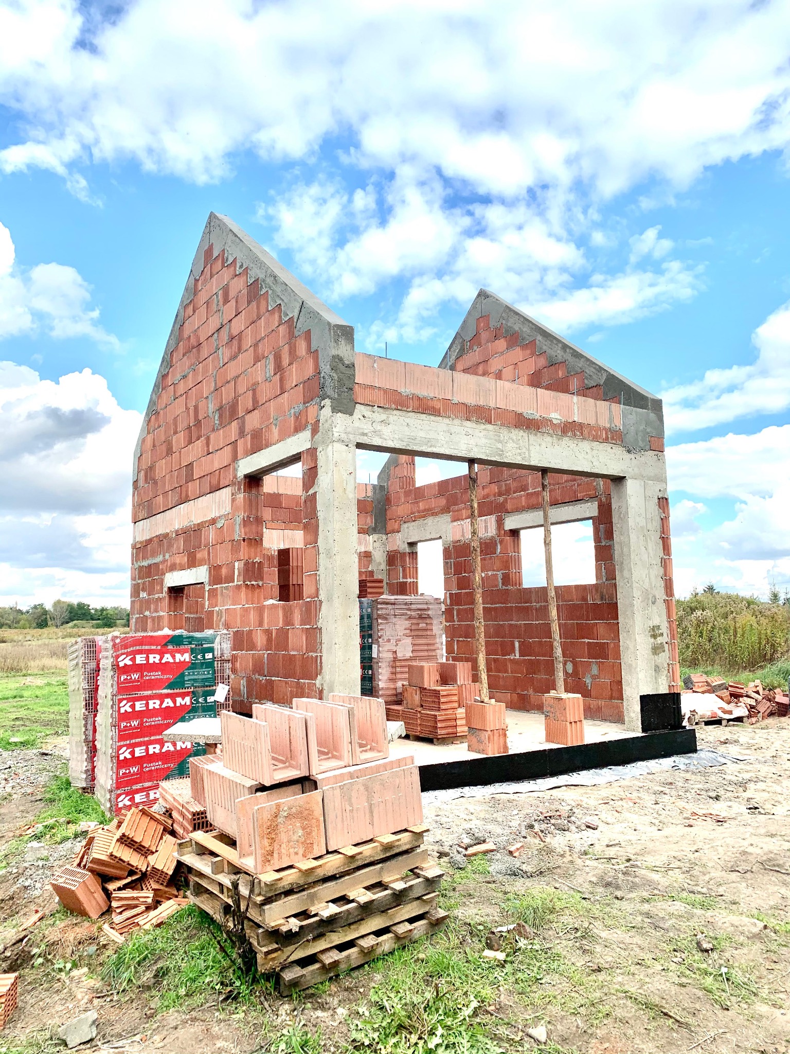 Wznoszenie ścian domu z czerwonej cegły ceramicznej, widoczne palety z cegłami, niebo z chmurami w tle. Budowa w toku, stan surowy otwarty.