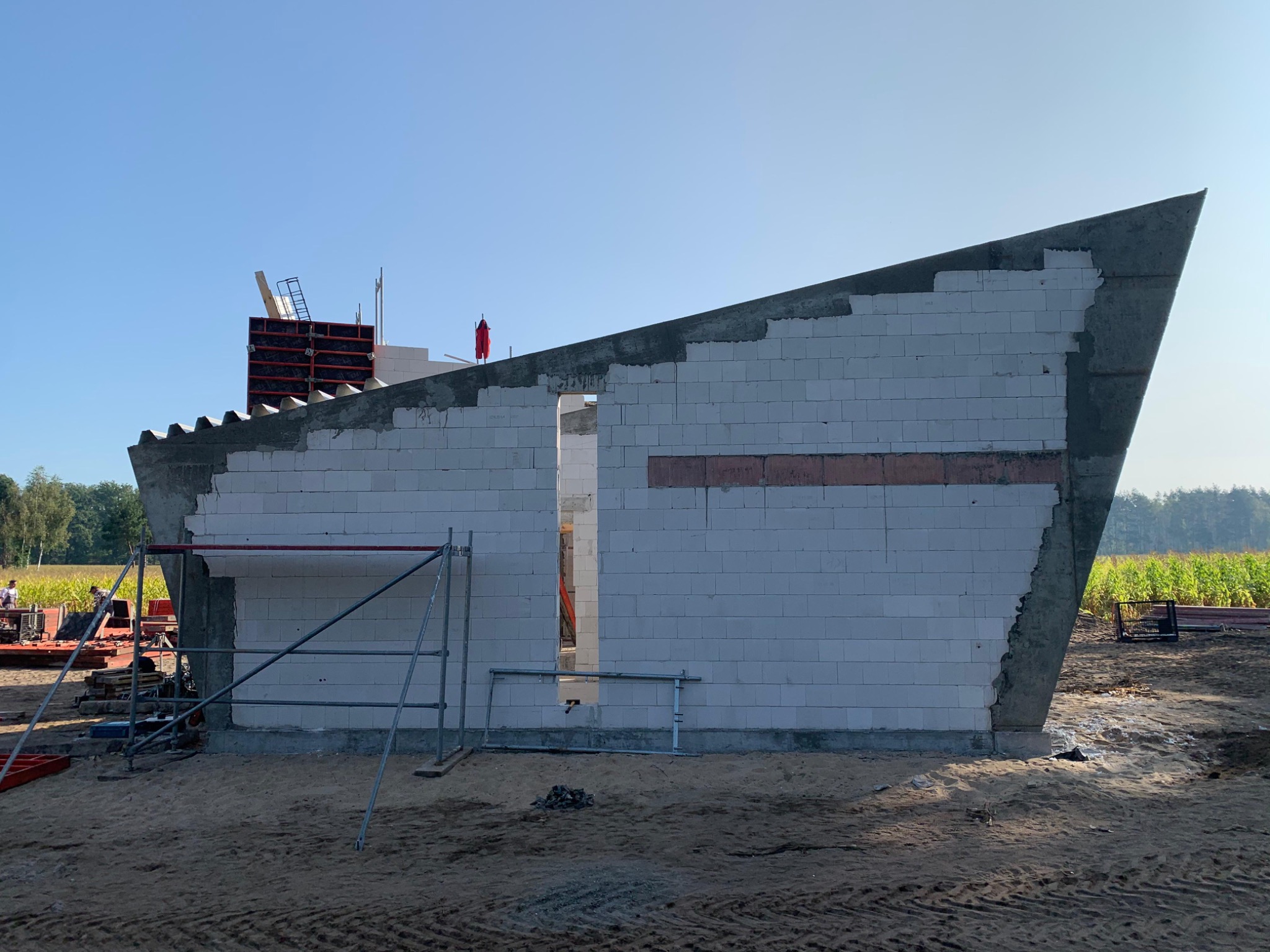 Nietypowy dom w budowie: białe bloczki, skośny dach, szalunki i rusztowania. Widok na konstrukcję w surowym stanie, z polami w tle.