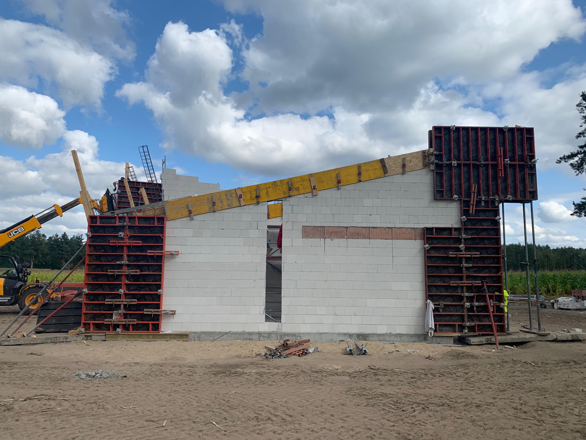 Wznoszenie ścian domu z białych bloczków, z użyciem szalunków systemowych i dźwigu teleskopowego JCB na tle błękitnego nieba z chmurami.