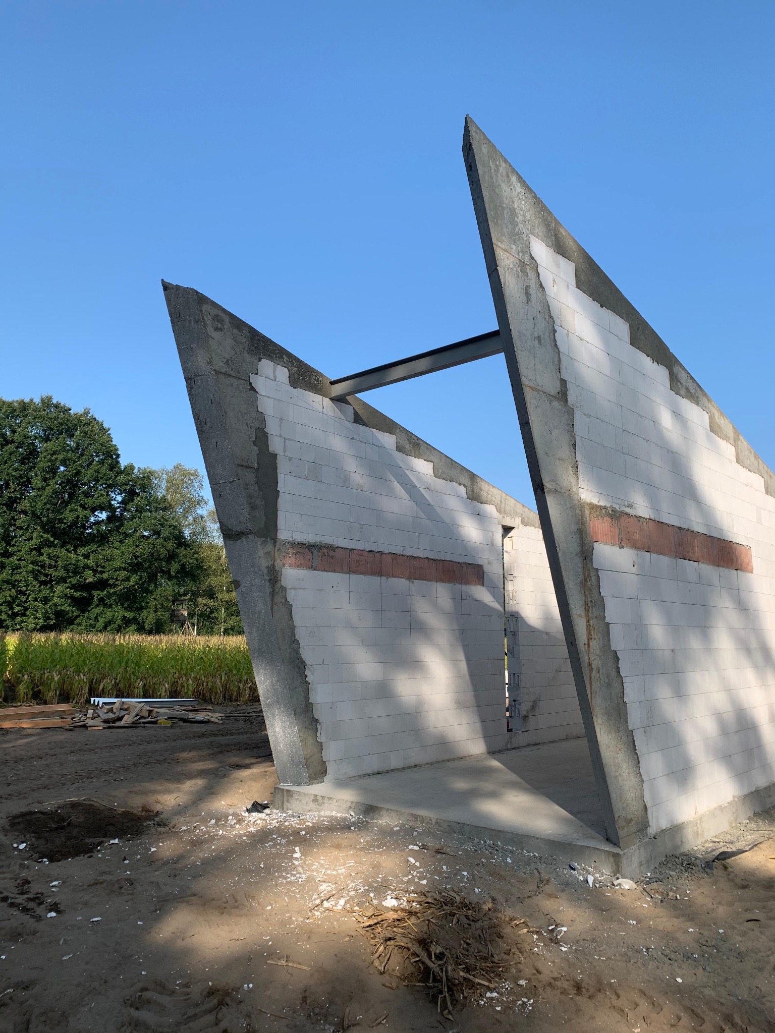 Surowe ściany z betonu komórkowego i żelbetu w nietypowym kształcie, z widoczną stalową belką, na tle błękitnego nieba i zieleni. Budowa w toku, widoczne materiały budowlane.