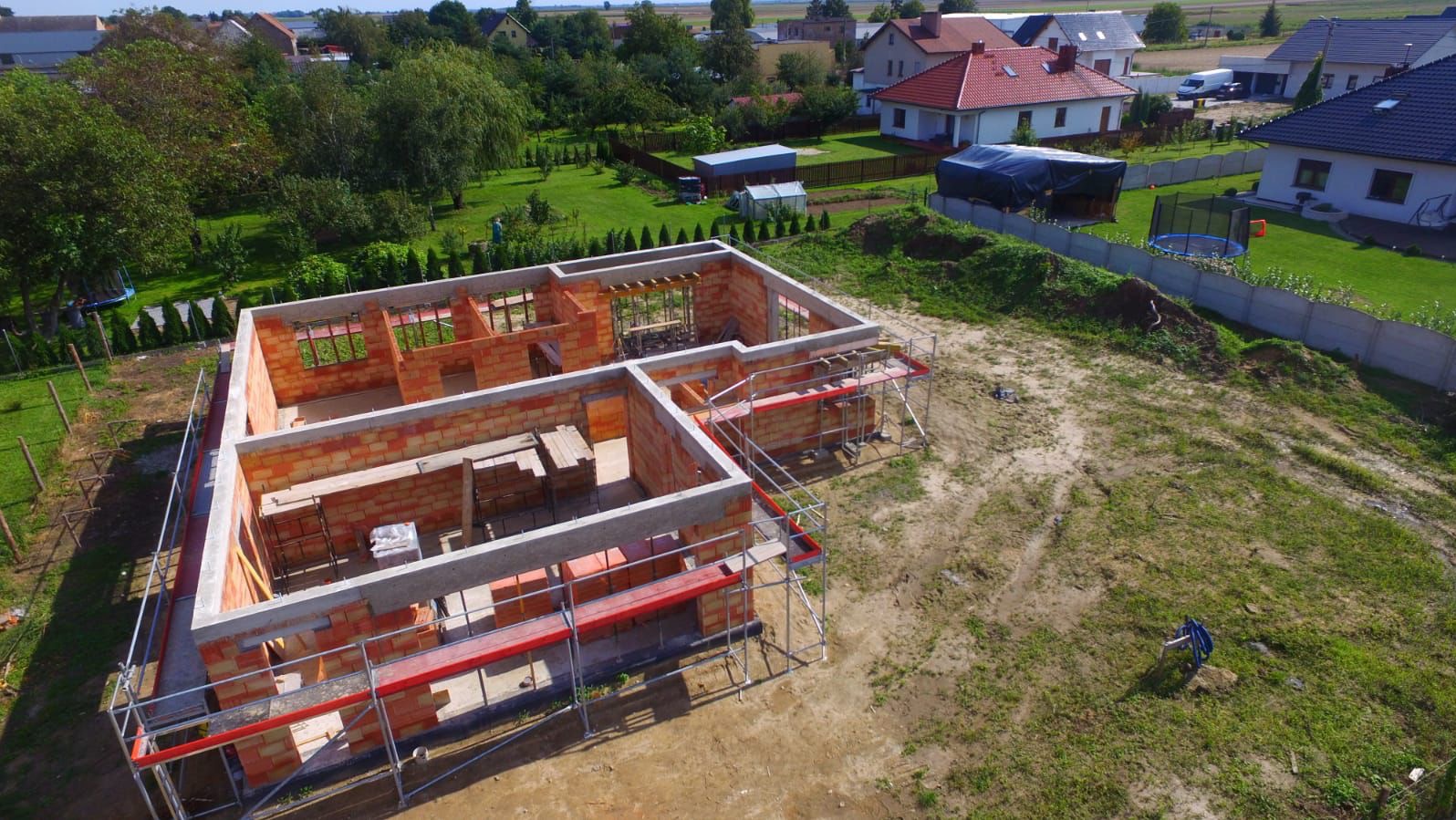 Wysoka perspektywa na budowę domu z czerwonej cegły, z widocznymi rusztowaniami i betonowymi nadprożami, na tle zielonego krajobrazu z domami.