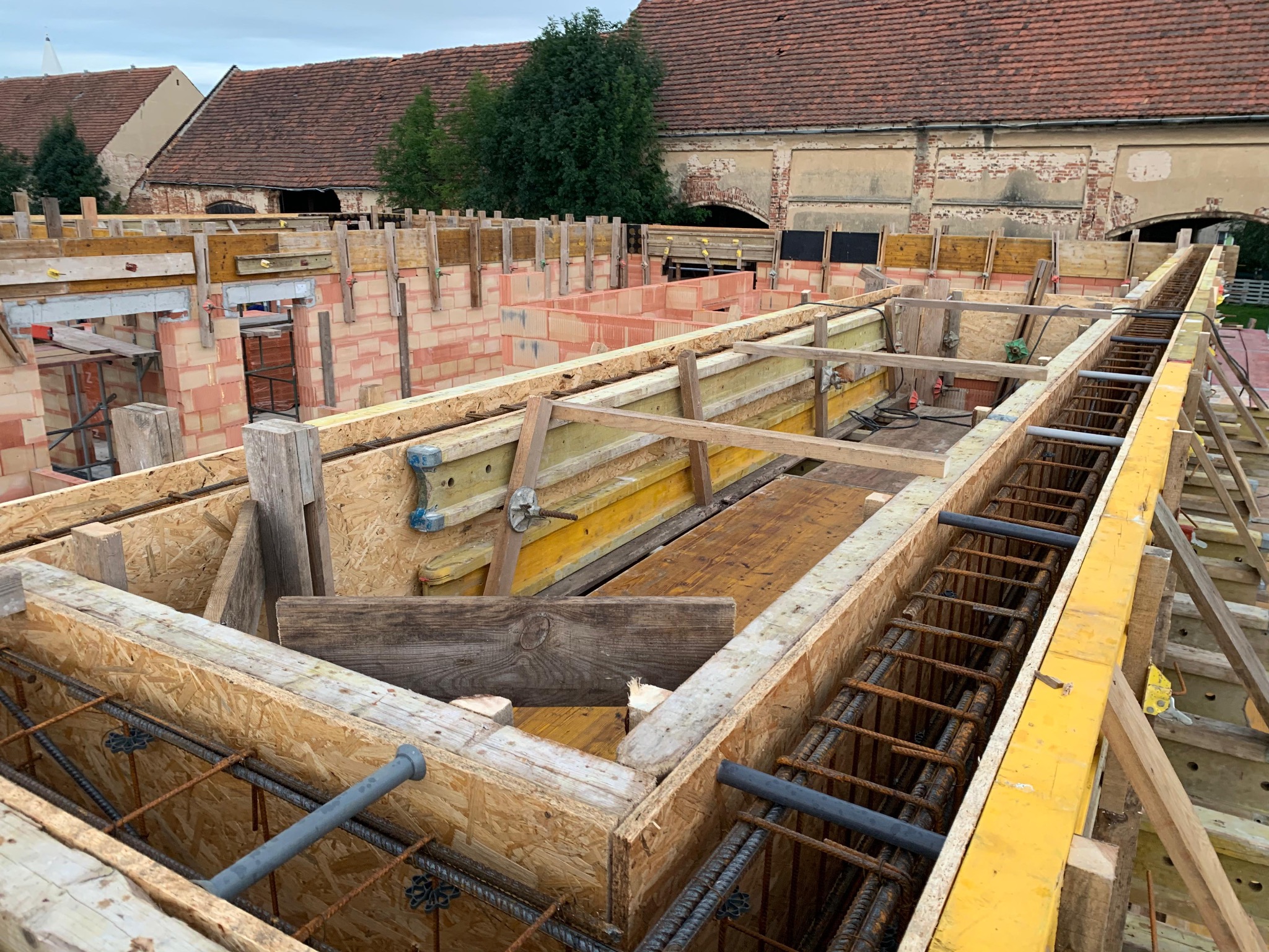 Zbrojenie fundamentów w drewnianym szalunku, widok z góry na plac budowy z ceglanymi budynkami w tle. Elementy stalowe i drewniane konstrukcji.