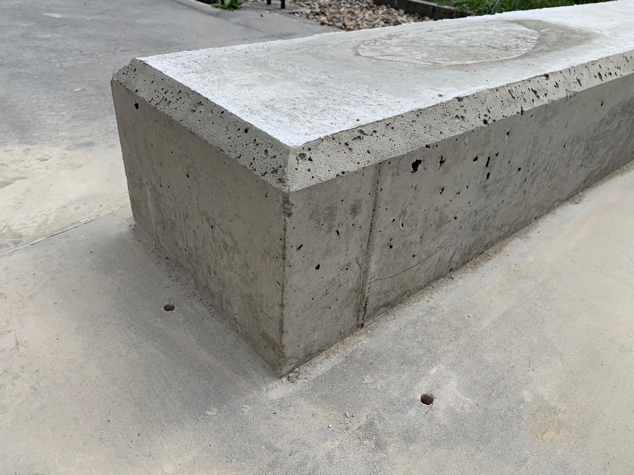 Betonowy element architektoniczny z fazowanymi krawędziami, widoczne pory w strukturze betonu, osadzony na betonowej powierzchni z otworami drenażowymi.