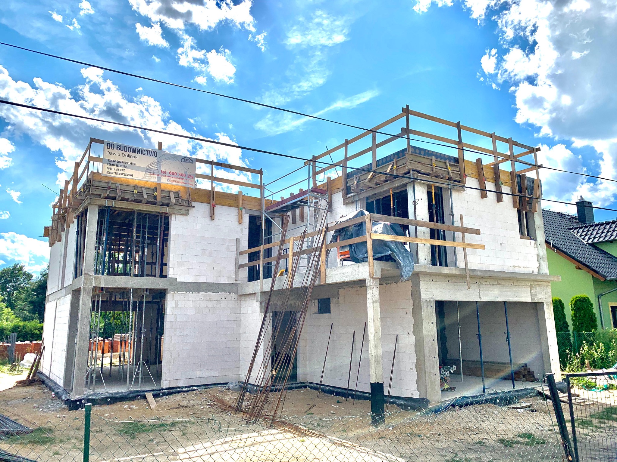 Wznoszenie dwukondygnacyjnego domu z białych bloczków, z widocznym zbrojeniem i drewnianymi barierkami. Na górze baner reklamowy firmy budowlanej. Błękitne niebo w tle.