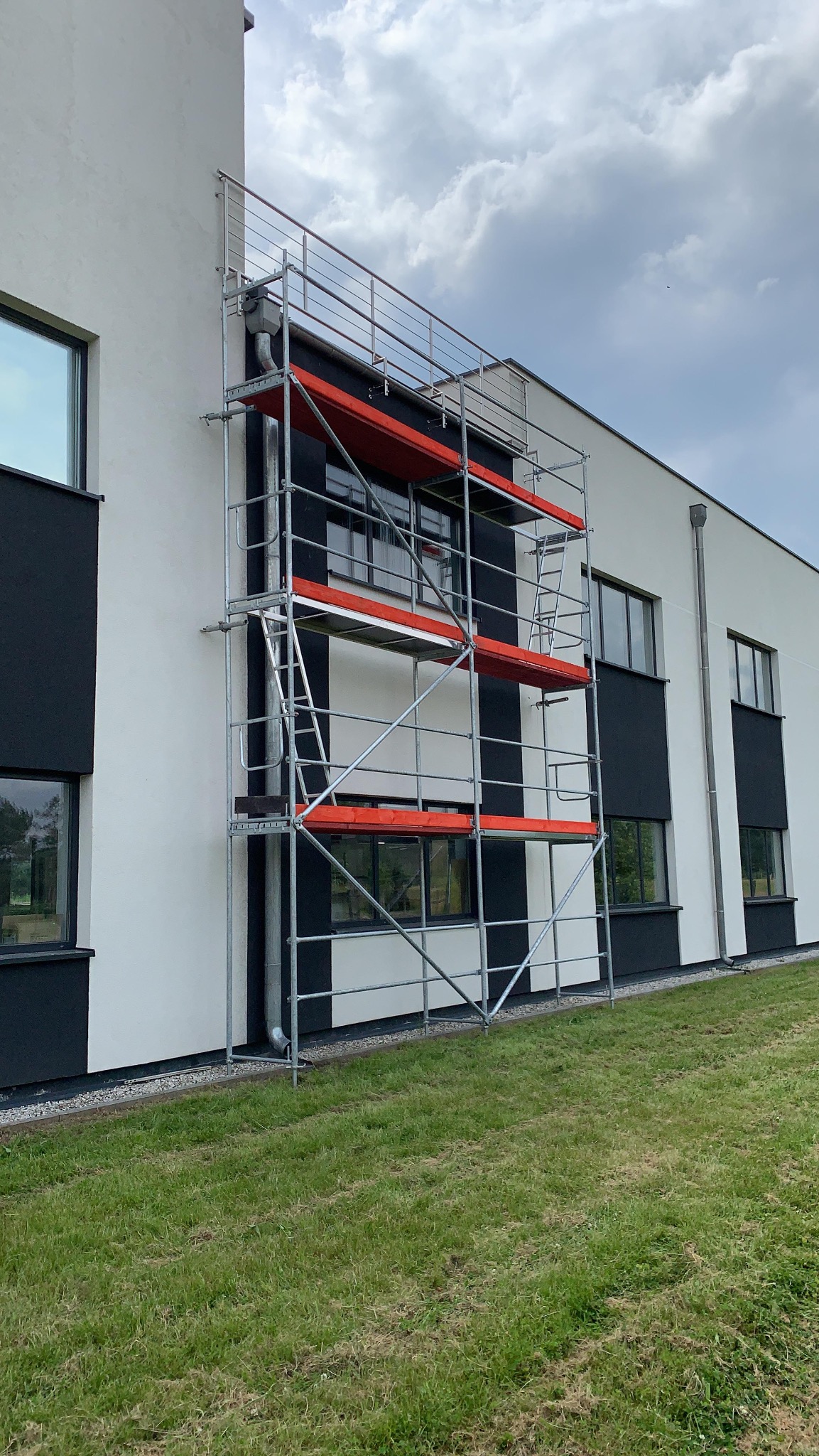 Elewacja budynku z rusztowaniem na tle zielonego trawnika i pochmurnego nieba. Widoczne okna i elementy wykończeniowe w kolorze czarnym.
