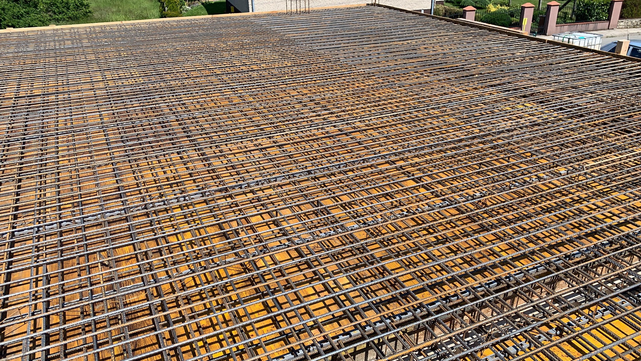 Siatka zbrojeniowa ułożona na deskowaniu, przygotowana do zalania betonem. Widok z góry, ukazujący regularny układ prętów i drewniane podłoże. Element konstrukcyjny budynku.