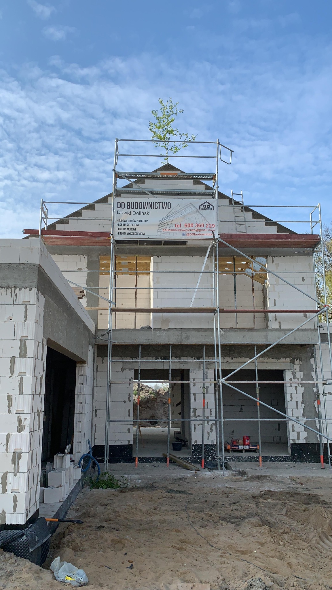 Budowa domu z białych bloczków, widoczne rusztowania i baner firmy DD Budownictwo. Częściowo otynkowane ściany, okna bez szyb. W tle błękitne niebo.