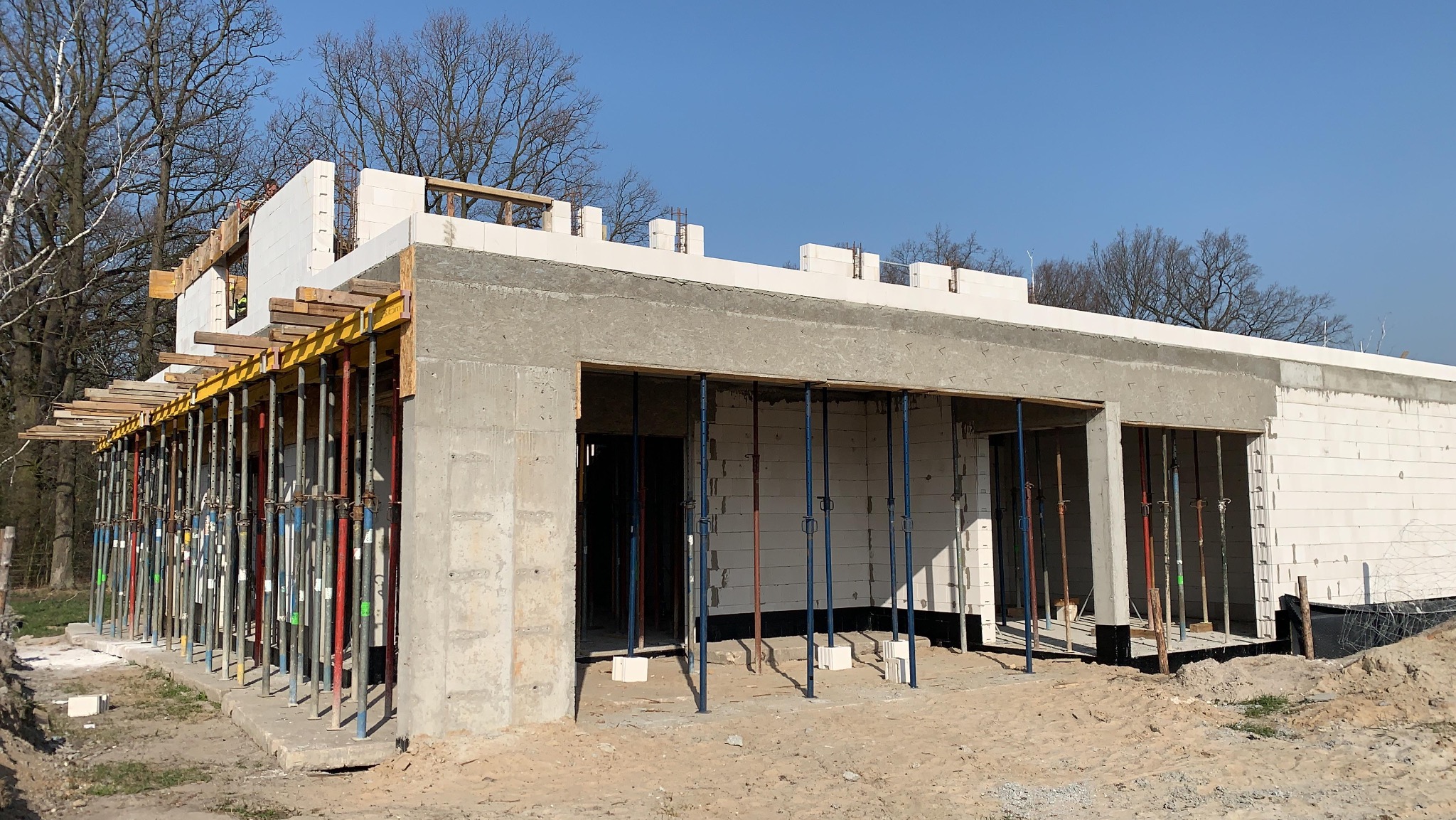 Budowa domu z betonu komórkowego w trakcie realizacji. Widoczne szalunki i podpory. Częściowo otynkowane ściany. W tle drzewa i błękitne niebo.