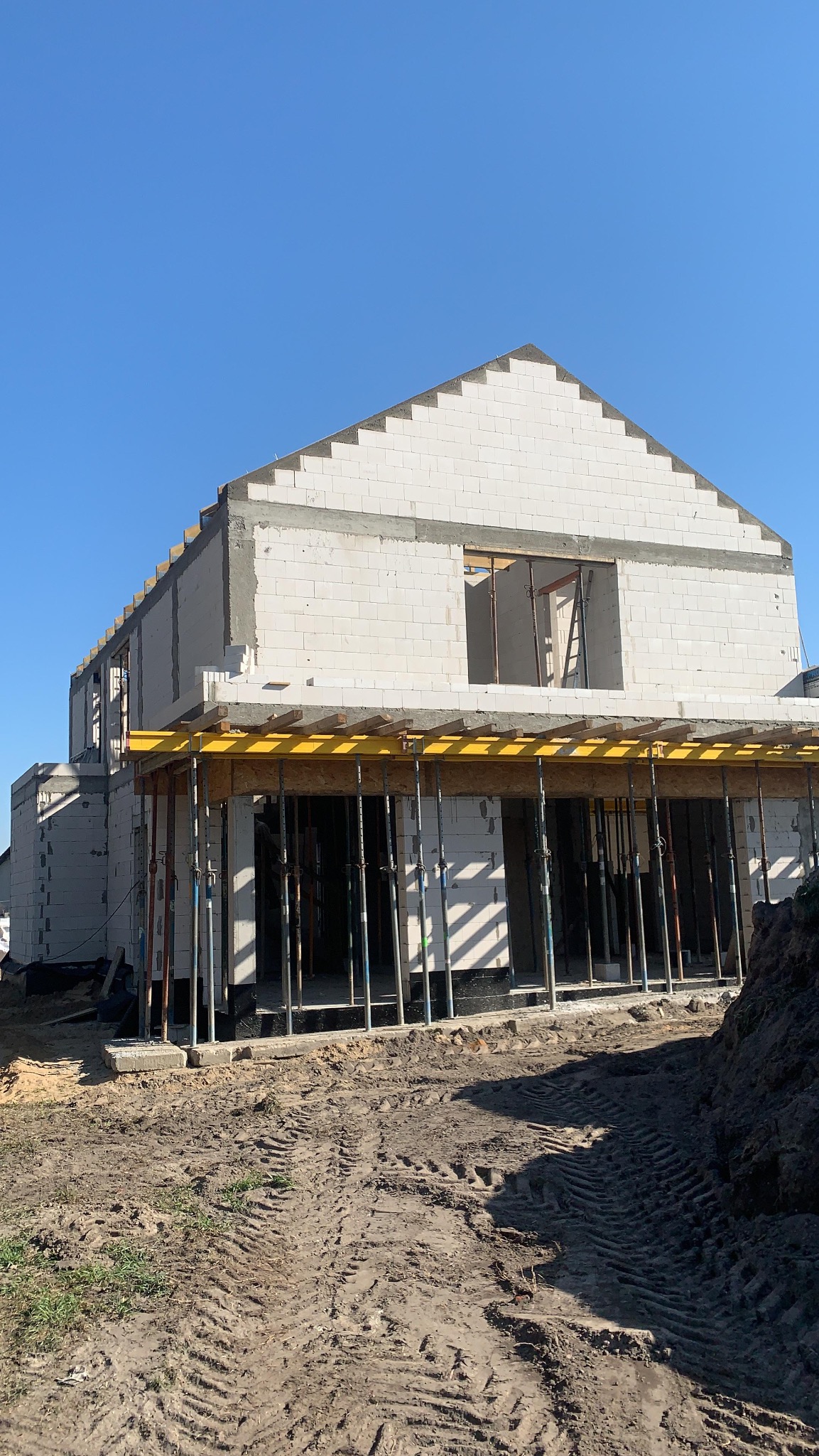 Wznoszenie ścian z białych bloczków na budowie domu jednorodzinnego. Widoczne podpory stropu i ślady opon na ziemi. Słoneczny dzień, błękitne niebo.