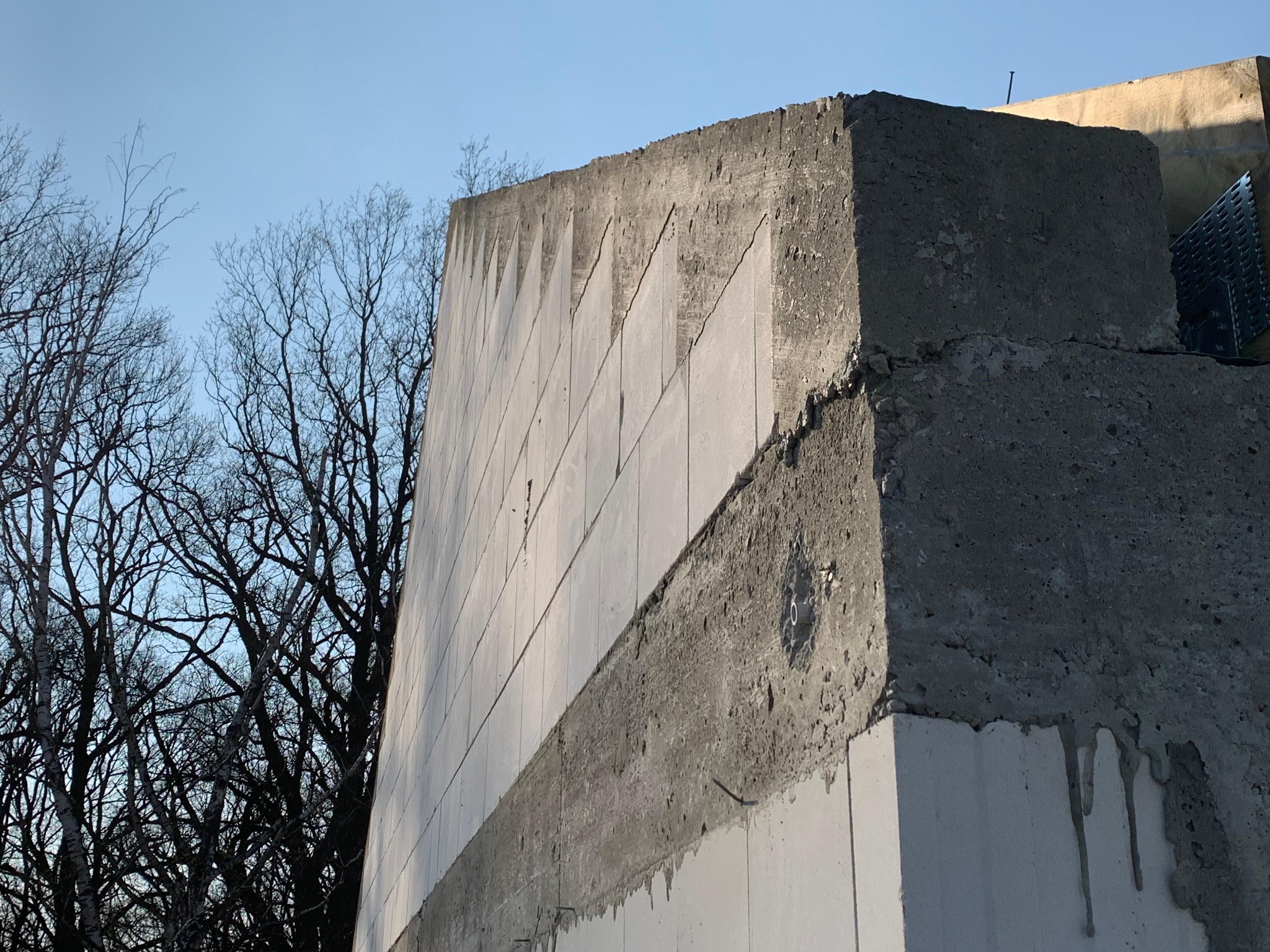 Surowa ściana z betonu komórkowego w trakcie budowy, widoczne elementy konstrukcyjne i fragmenty drzew na tle jasnego nieba. Detal budowlany w zbliżeniu.