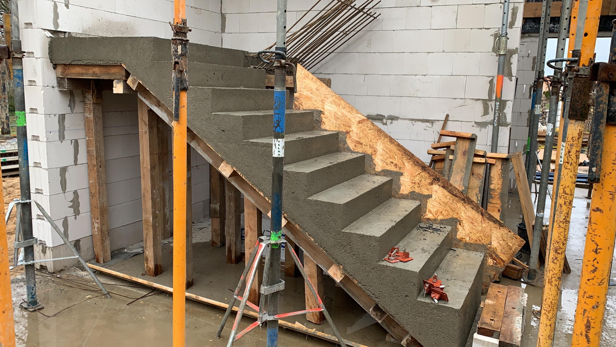 Betonowe schody w trakcie budowy, widoczne szalunki i podpory. Ściany z bloczków betonowych, zbrojenie w tle. Etap wylewania betonu na schody.