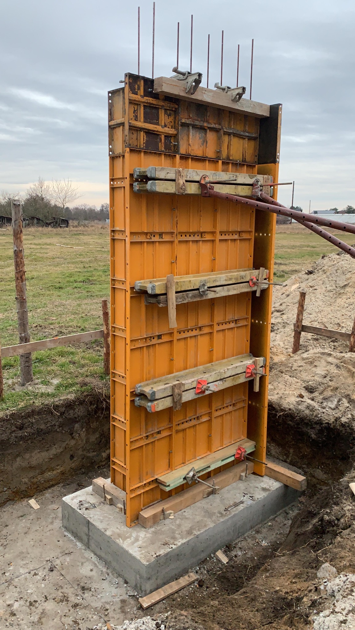 Żółte szalunki kolumny na fundamencie z wystającymi prętami zbrojeniowymi, widoczne drewniane podpory i ziemia wokół, pochmurne niebo w tle.