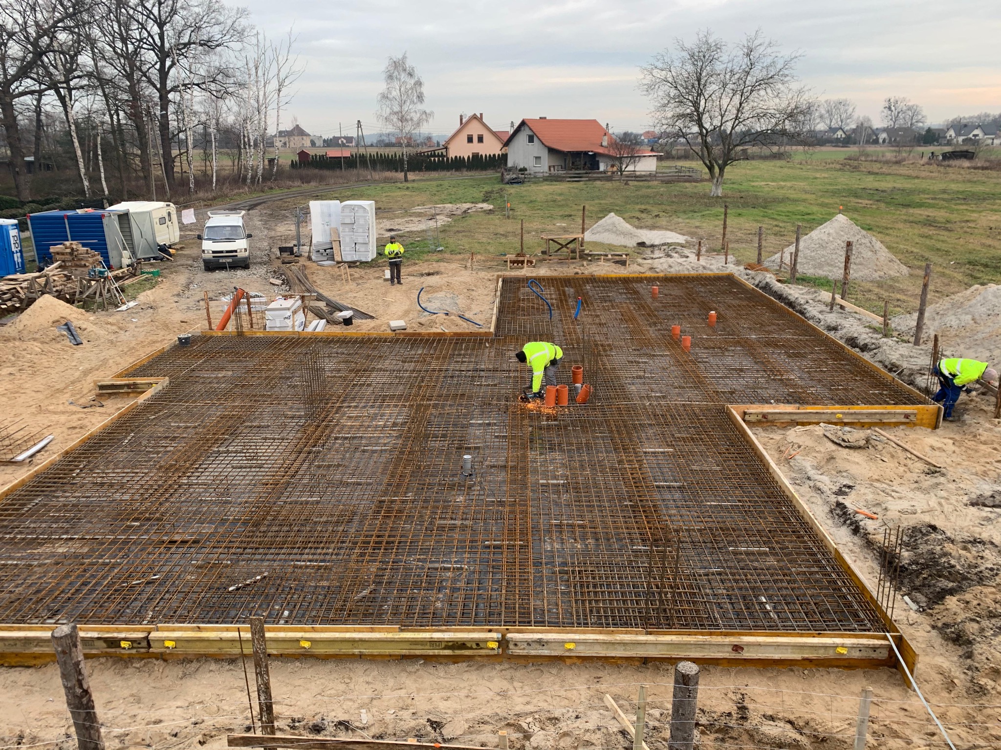 Zbrojenie fundamentu pod dom jednorodzinny z widocznymi pracownikami w odblaskowych kamizelkach i elementami szalunku. W tle zabudowa wiejska i drzewa.