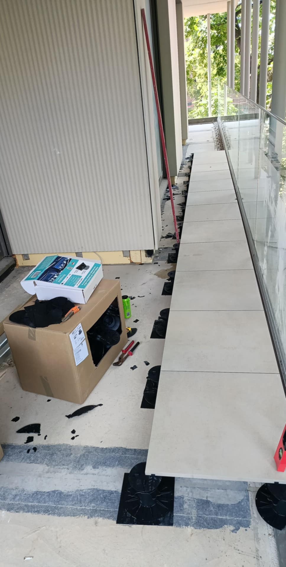 Balkon w trakcie układania płytek na specjalnych podkładkach dystansowych. Widoczny karton z akcesoriami, poziomica i narzędzia. Praca w toku na zewnątrz budynku.